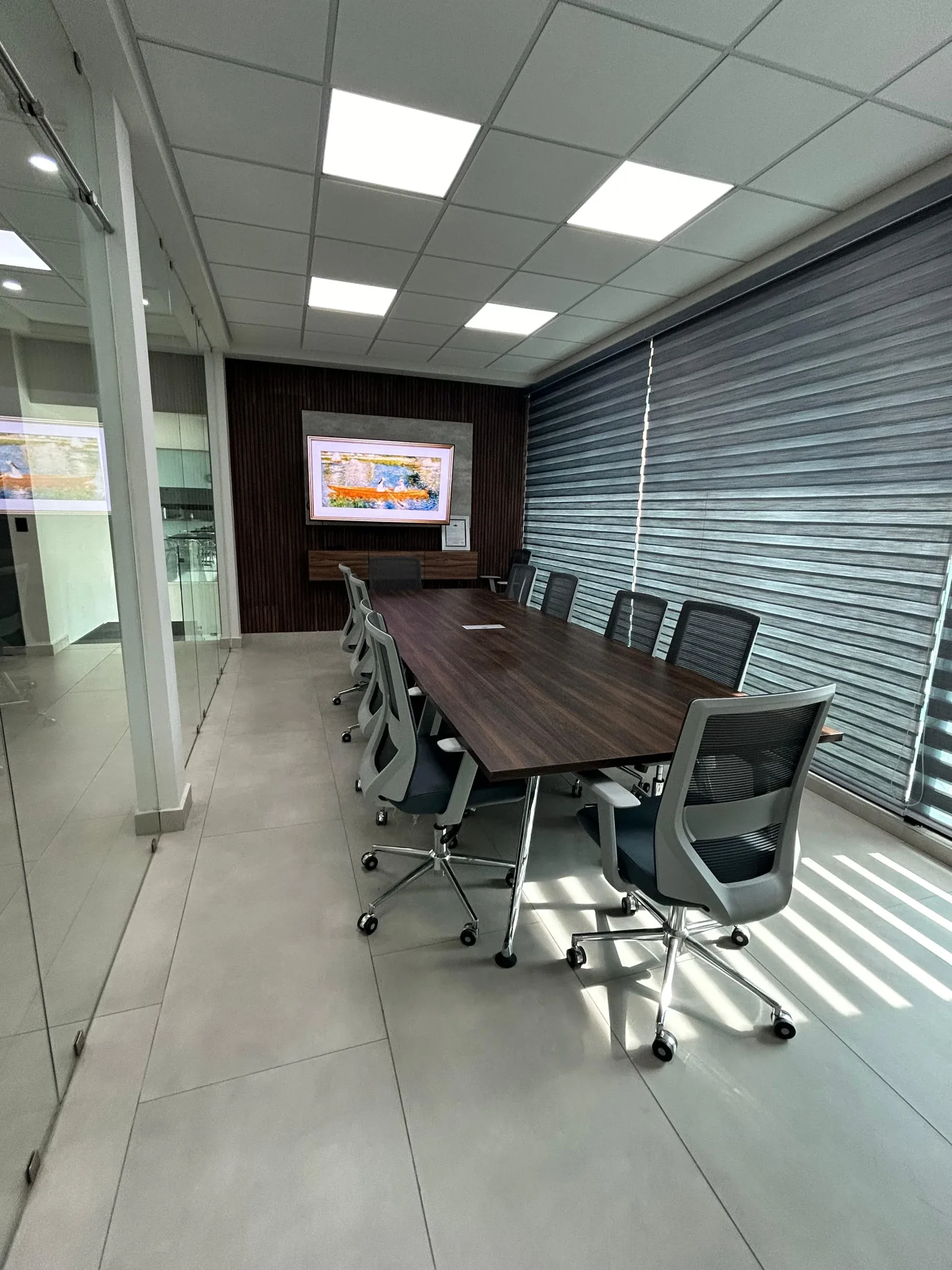 Sala de juntas con mesa de madera oscura, sillas de malla y pantalla sobre panel ranurado