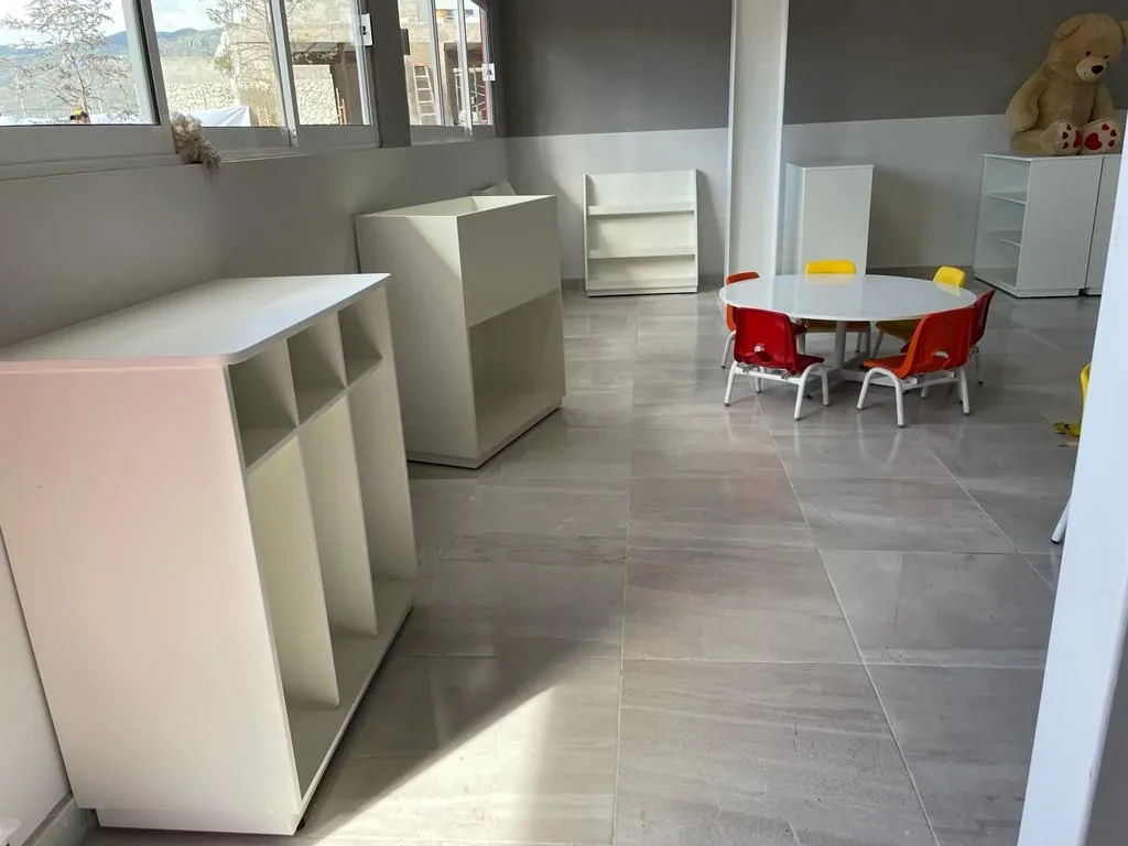 Aula infantil equipada con muebles bajos de melamina blanca, mesa redonda y sillas de colores