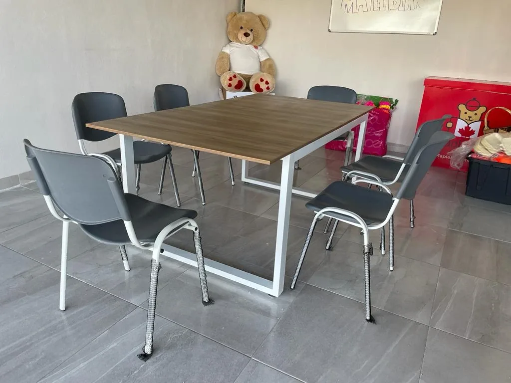 Mesa rectangular con cubierta de madera y estructura metálica blanca, con sillas apilables